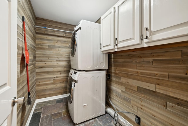 laundry room featuring stacked washing maching and dryer, cabinet space, wood walls, and stone tile flooring