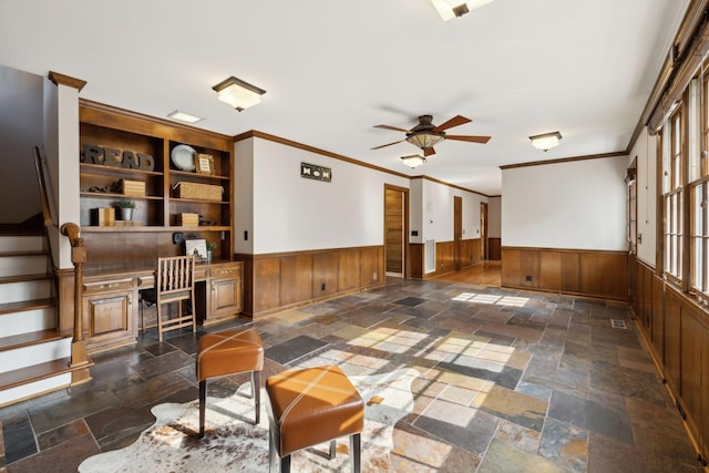 interior space featuring crown molding, a ceiling fan, a wainscoted wall, and stone tile flooring