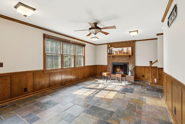 unfurnished living room with stone tile flooring, ornamental molding, a brick fireplace, a wainscoted wall, and a ceiling fan
