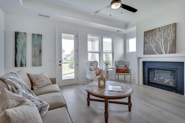 living area with visible vents, ornamental molding, a glass covered fireplace, ceiling fan, and light wood finished floors