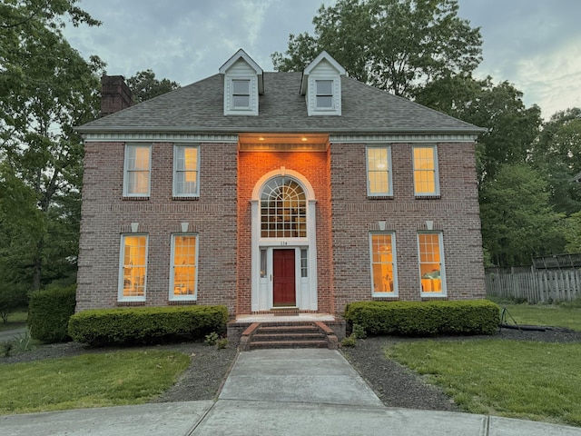colonial house with fence, a front yard, roof with shingles, a chimney, and brick siding
