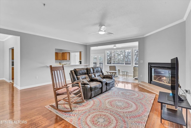 living room featuring ceiling fan, light wood-style floors, a glass covered fireplace, baseboards, and ornamental molding