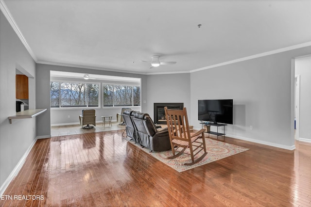 living room with ceiling fan, baseboards, wood-type flooring, a glass covered fireplace, and ornamental molding