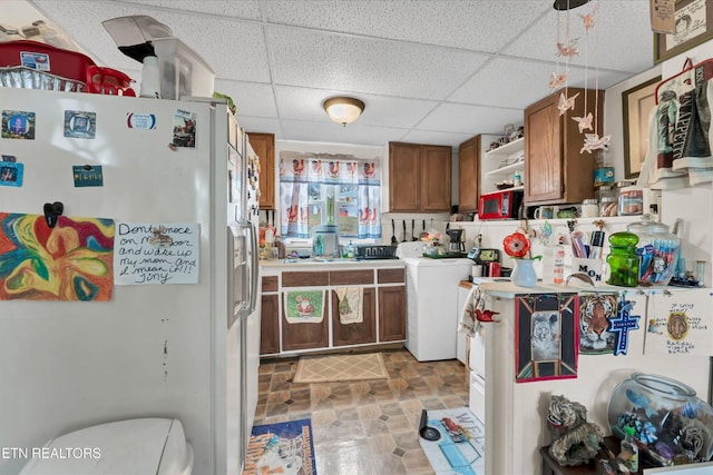 kitchen with open shelves, white refrigerator with ice dispenser, brown cabinetry, a paneled ceiling, and washer / dryer