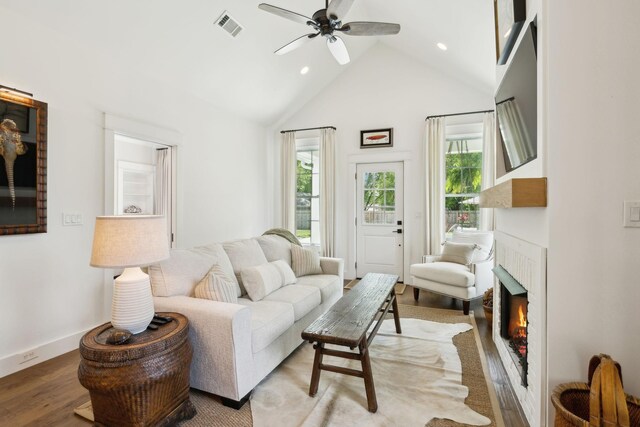 living area with ceiling fan, visible vents, wood finished floors, high vaulted ceiling, and a warm lit fireplace