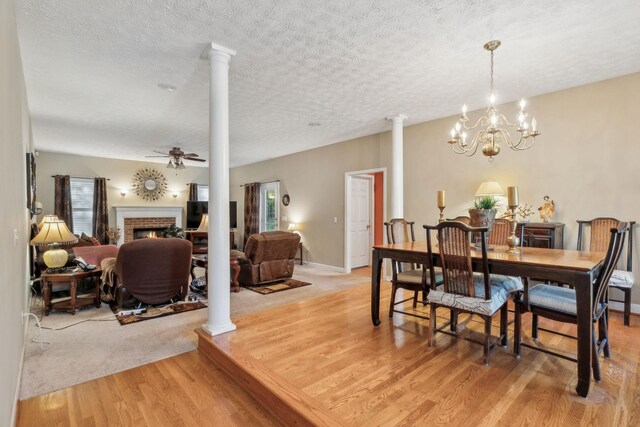 dining space with a fireplace, ornate columns, a textured ceiling, light wood finished floors, and ceiling fan with notable chandelier