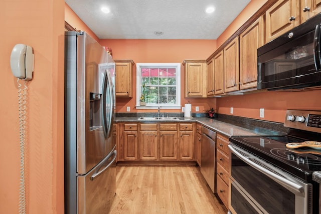 kitchen with dark countertops, appliances with stainless steel finishes, a sink, brown cabinetry, and light wood-style flooring