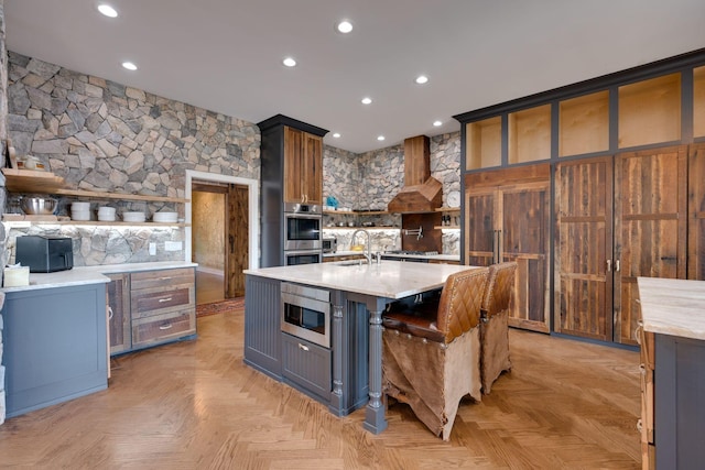 kitchen featuring open shelves, a center island with sink, wall chimney range hood, recessed lighting, and appliances with stainless steel finishes