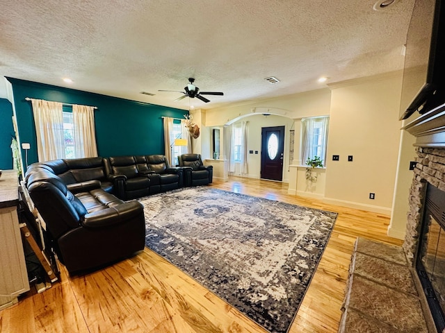 living room with a textured ceiling, a fireplace, light wood-style flooring, arched walkways, and a ceiling fan