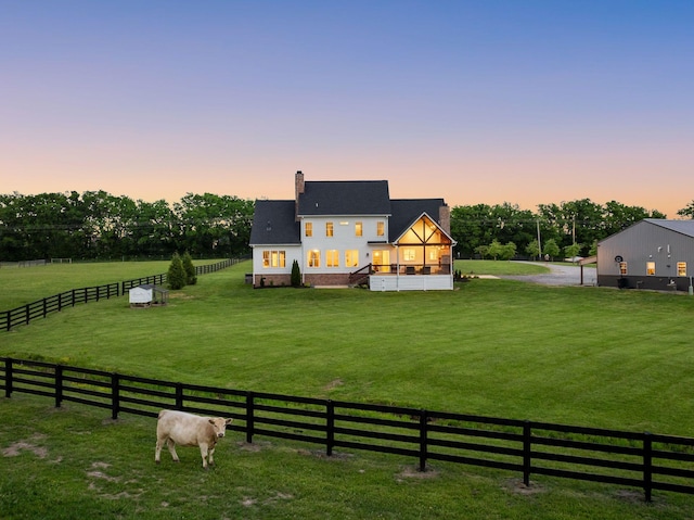 back of house at dusk with fence, a sunroom, a chimney, a rural view, and a lawn