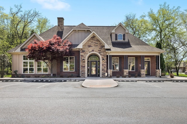 craftsman inspired home with roof with shingles, brick siding, board and batten siding, a chimney, and uncovered parking