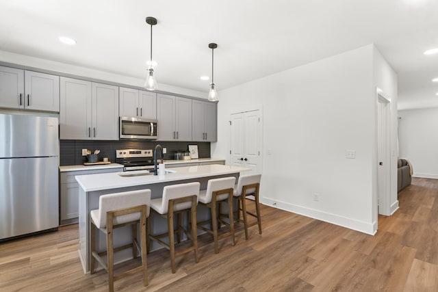 kitchen with an island with sink, light countertops, a sink, appliances with stainless steel finishes, and gray cabinetry