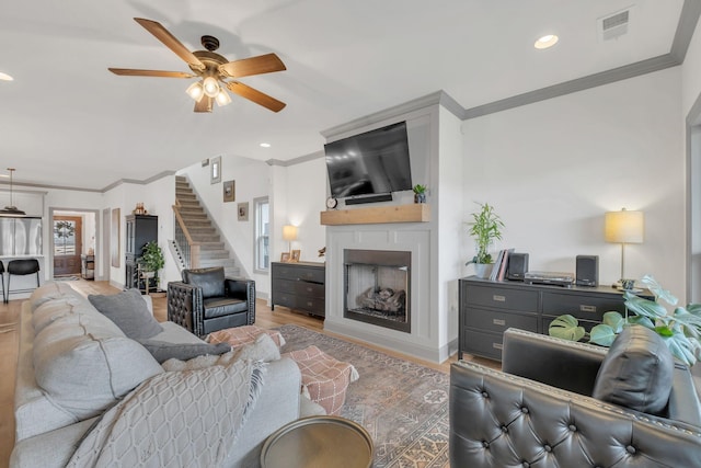 living area with visible vents, ornamental molding, stairs, a large fireplace, and a ceiling fan
