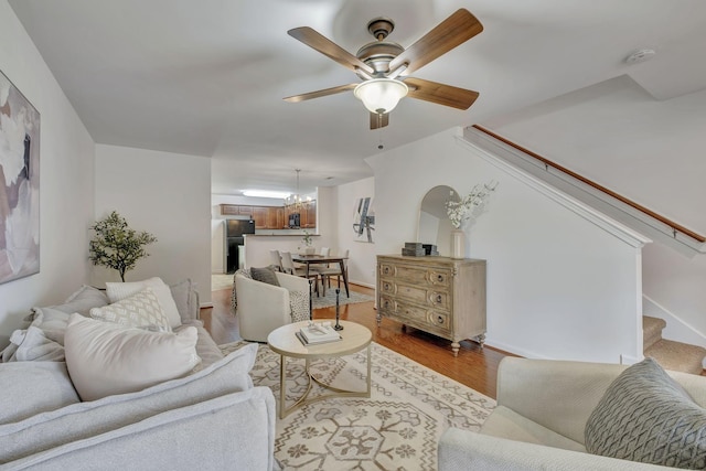 living room featuring stairway, baseboards, light wood-style floors, and ceiling fan with notable chandelier