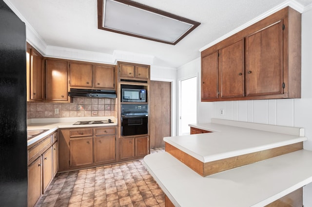 kitchen with brown cabinets, light countertops, black appliances, a peninsula, and under cabinet range hood