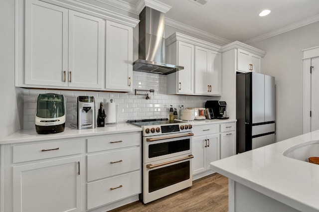 kitchen with wall chimney range hood, freestanding refrigerator, range with two ovens, white cabinetry, and ornamental molding
