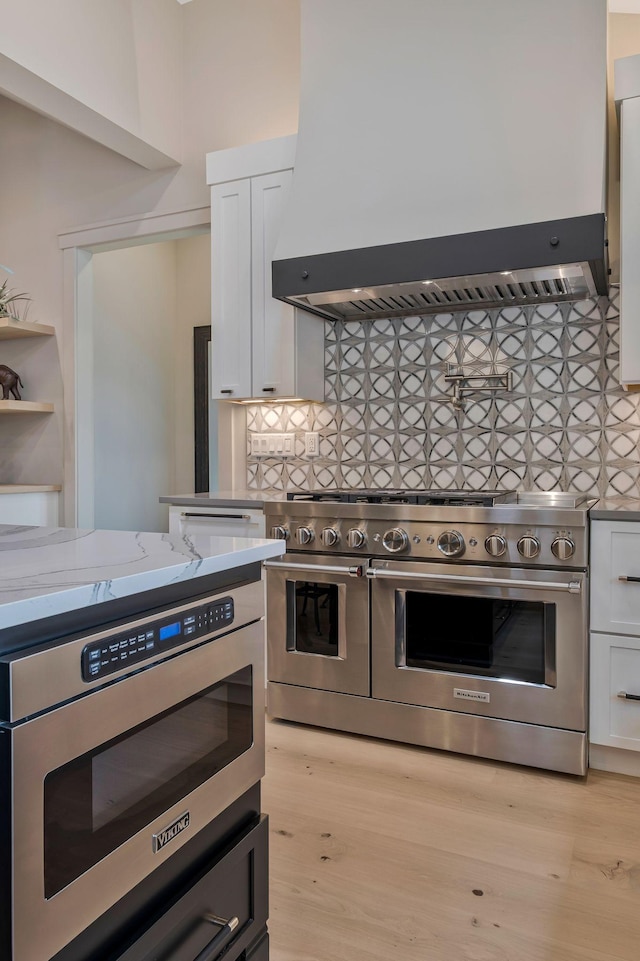 kitchen with backsplash, appliances with stainless steel finishes, white cabinetry, light stone counters, and wall chimney exhaust hood