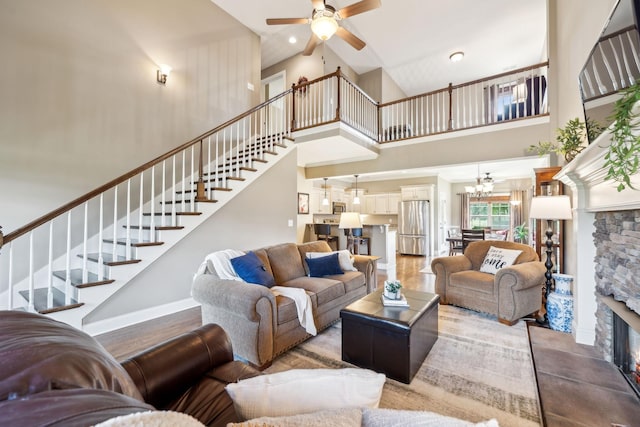 living room with stairs, a towering ceiling, a stone fireplace, baseboards, and ceiling fan with notable chandelier