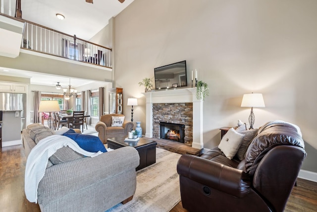 living area featuring a fireplace, ceiling fan with notable chandelier, baseboards, a towering ceiling, and dark wood-type flooring