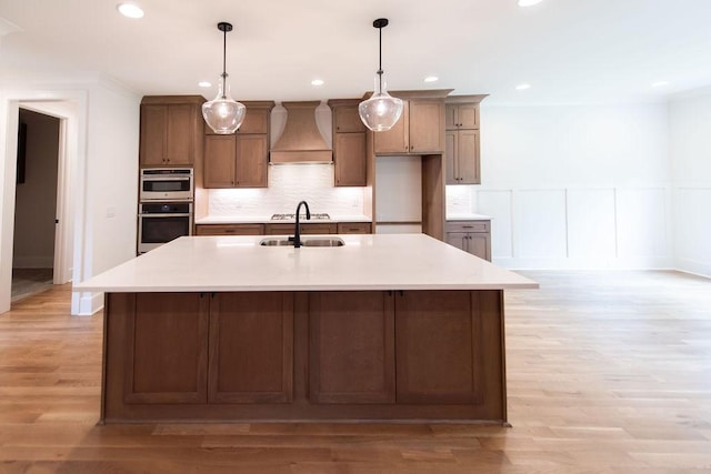 kitchen featuring decorative backsplash, a kitchen island with sink, double oven, premium range hood, and a sink