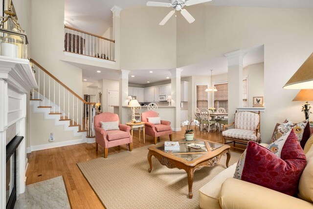 living area featuring a fireplace, ceiling fan with notable chandelier, light wood-type flooring, a towering ceiling, and ornate columns