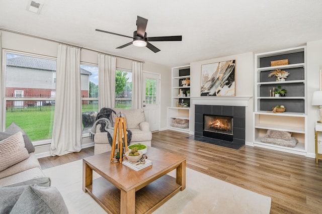 living area with a tiled fireplace, built in shelves, ceiling fan, and wood finished floors