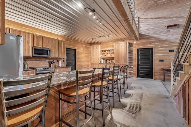 kitchen with concrete flooring, a breakfast bar, wooden walls, rail lighting, and appliances with stainless steel finishes