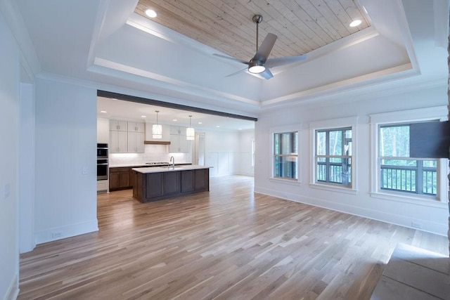 unfurnished living room featuring a ceiling fan, a tray ceiling, a sink, and light wood-style flooring