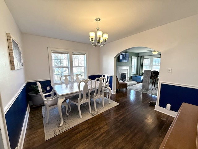 dining area featuring arched walkways, a brick fireplace, wood finished floors, and ceiling fan with notable chandelier