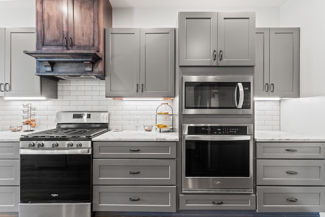kitchen featuring decorative backsplash, light stone countertops, custom range hood, appliances with stainless steel finishes, and gray cabinetry