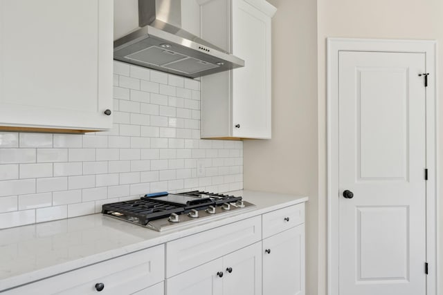 kitchen with wall chimney range hood, white cabinets, light stone counters, stainless steel gas stovetop, and backsplash