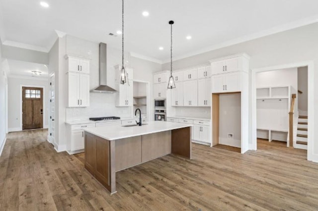 kitchen featuring wall chimney range hood, light countertops, ornamental molding, and an island with sink