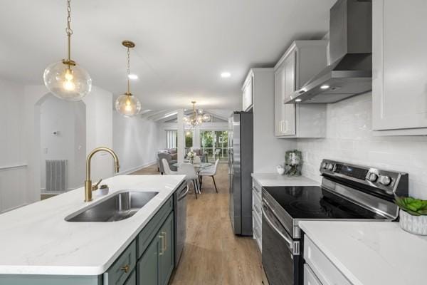kitchen featuring an island with sink, wall chimney exhaust hood, a sink, light wood finished floors, and appliances with stainless steel finishes