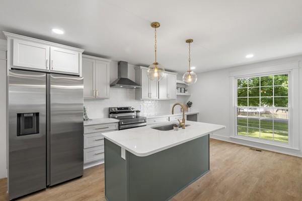 kitchen featuring a kitchen island with sink, wall chimney range hood, light countertops, a sink, and appliances with stainless steel finishes