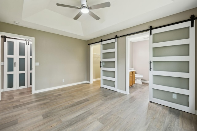 unfurnished bedroom featuring a barn door, light wood finished floors, ensuite bath, baseboards, and a tray ceiling