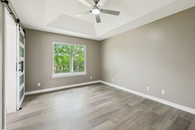empty room with baseboards, a barn door, a raised ceiling, a ceiling fan, and light wood finished floors