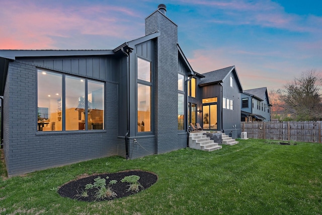 back of property at dusk featuring board and batten siding, a chimney, brick siding, fence, and a yard