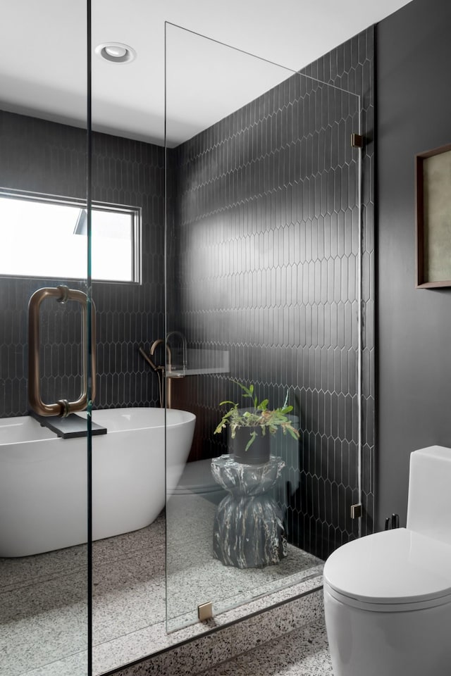 bathroom with speckled floor, a freestanding bath, toilet, and tile walls
