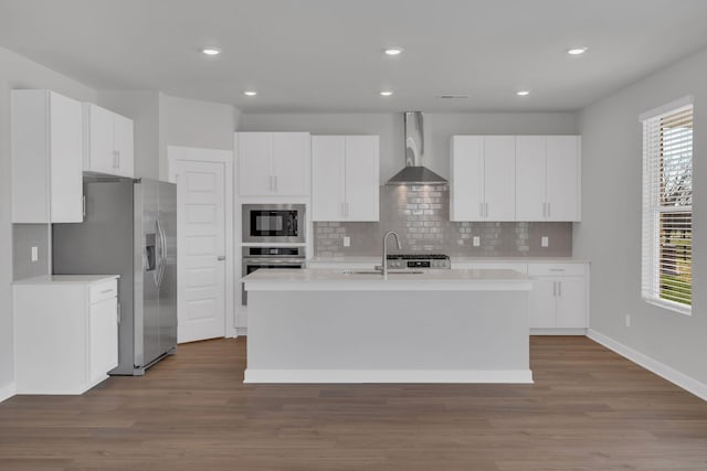 kitchen with a kitchen island with sink, wall chimney range hood, a sink, appliances with stainless steel finishes, and backsplash