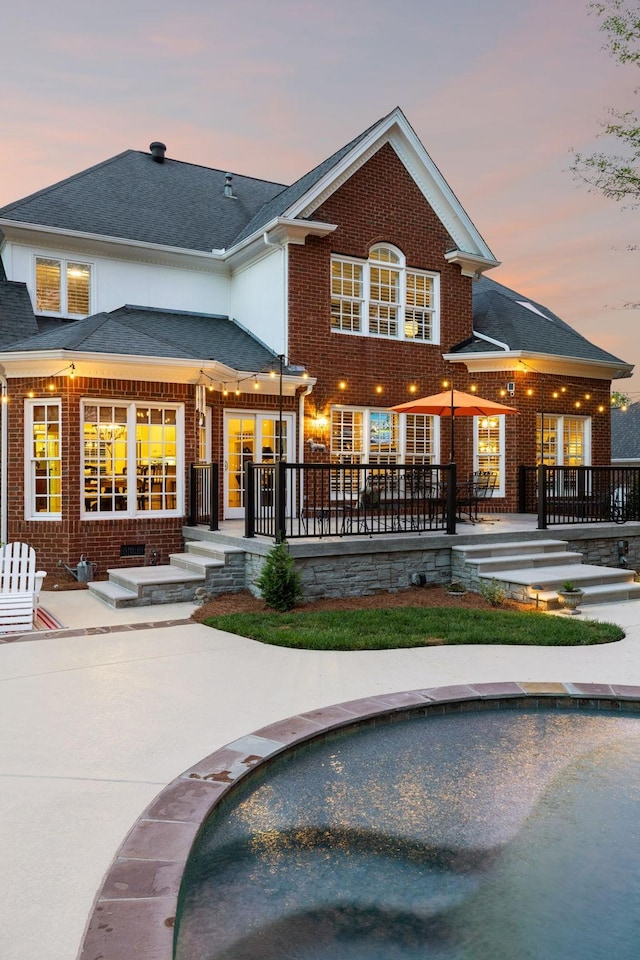 back of property at dusk with a patio, a shingled roof, an outdoor pool, and brick siding