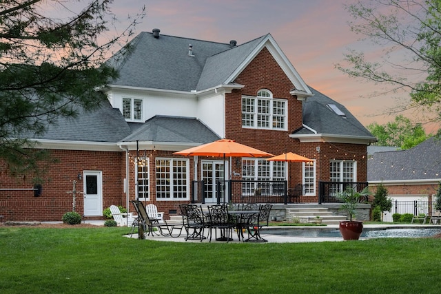 back of house at dusk featuring a lawn, brick siding, a shingled roof, and a patio area