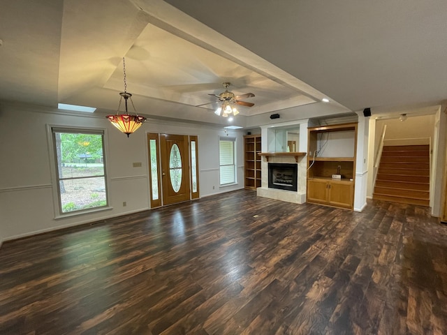 unfurnished living room with dark wood-style flooring, stairway, a fireplace with raised hearth, a tray ceiling, and a ceiling fan