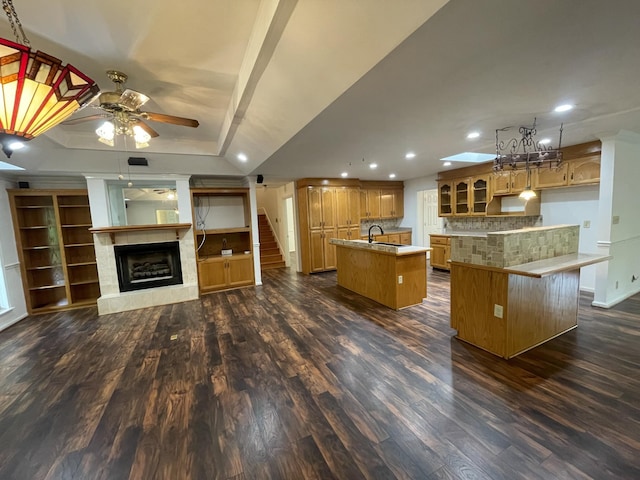 kitchen featuring ceiling fan, a center island with sink, a sink, a raised ceiling, and dark wood-type flooring