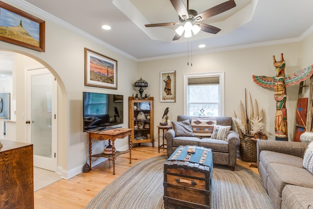 living area with crown molding, ceiling fan, arched walkways, light wood-type flooring, and a tray ceiling