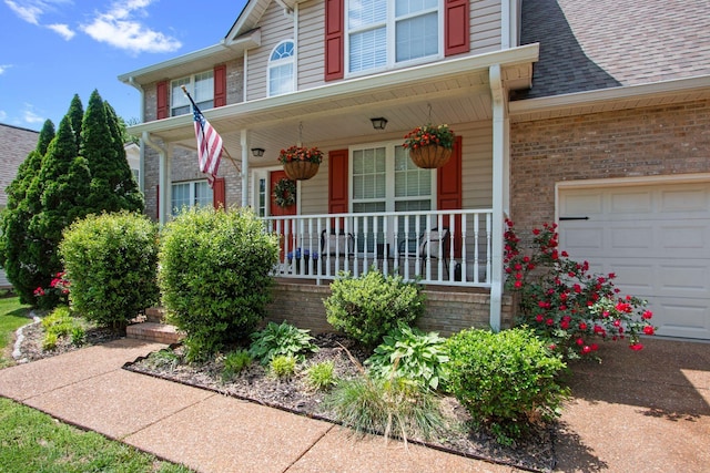 doorway to property featuring a porch, brick siding, an attached garage, and a shingled roof