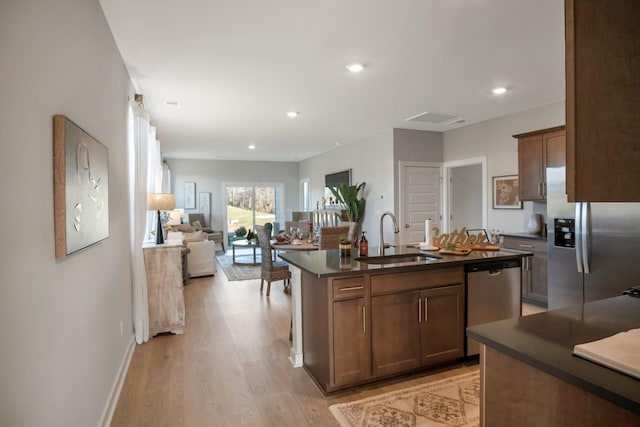 kitchen featuring appliances with stainless steel finishes, dark countertops, a sink, light wood finished floors, and an island with sink