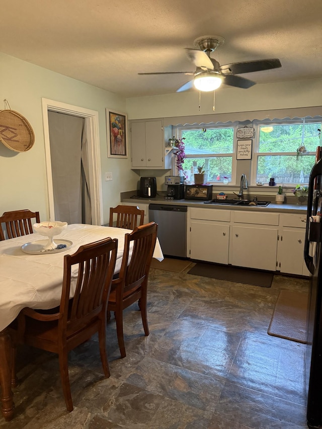 kitchen with stainless steel dishwasher, a ceiling fan, dark countertops, white cabinets, and a sink