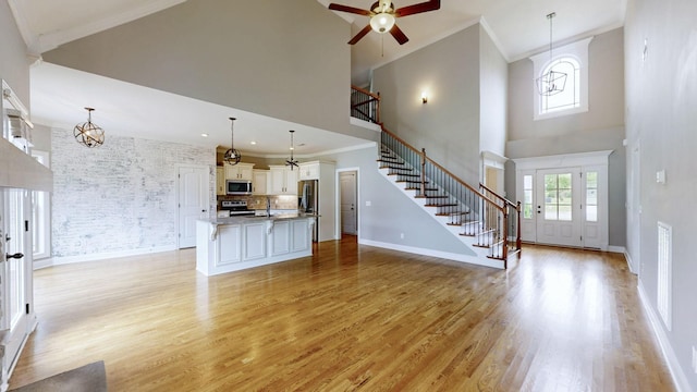 unfurnished living room featuring stairs, ceiling fan with notable chandelier, ornamental molding, a towering ceiling, and light wood-type flooring