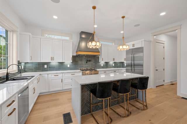 kitchen with stainless steel appliances, custom range hood, a sink, light wood finished floors, and backsplash