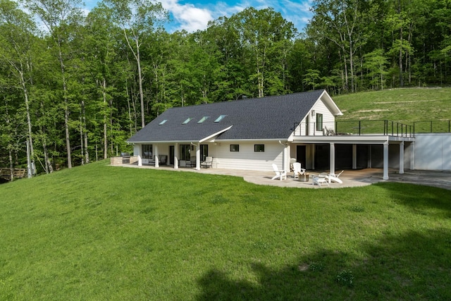 rear view of house with a patio, a shingled roof, a view of trees, and a lawn